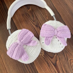 Puede incluir: Calentadores de orejas de crochet blanco con lazos morados. Los calentadores de orejas est&aacute;n unidos a una diadema blanca.