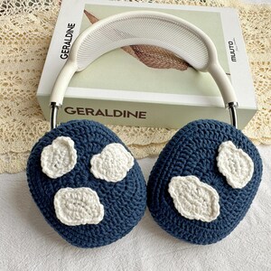 May include: Blue crocheted ear warmers with white crocheted cloud designs. The ear warmers are on a white book with the word "GERALDINE" printed on the cover.
