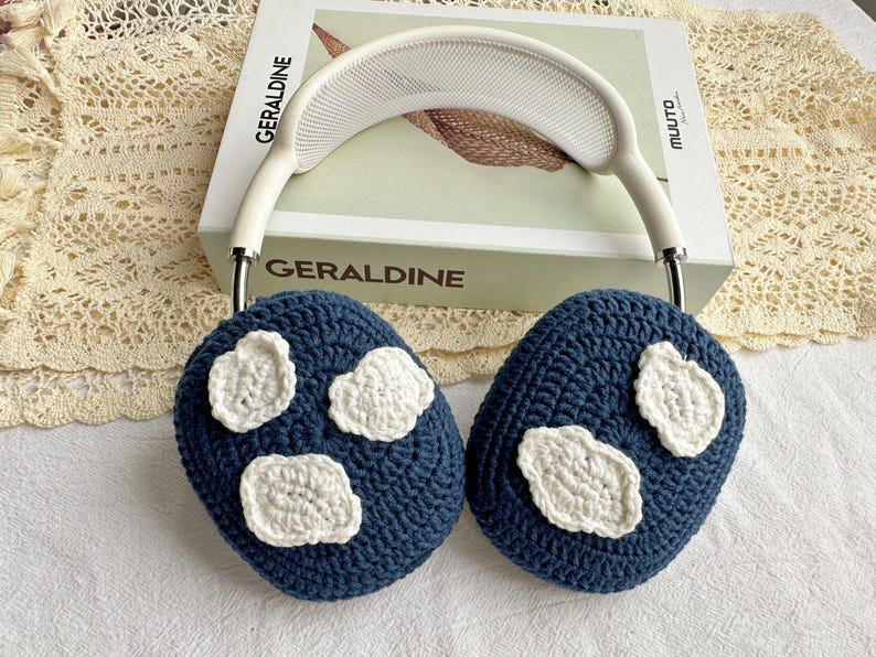 May include: Blue crocheted ear warmers with white crocheted cloud designs. The ear warmers are on a white book with the word "GERALDINE" printed on the cover.