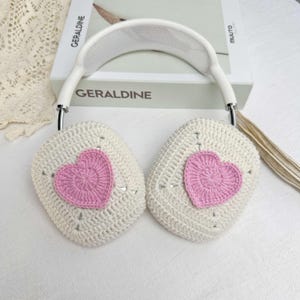 May include: White crocheted earmuffs with pink heart-shaped crochet embellishments. The earmuffs are attached to a white headband with a mesh design.