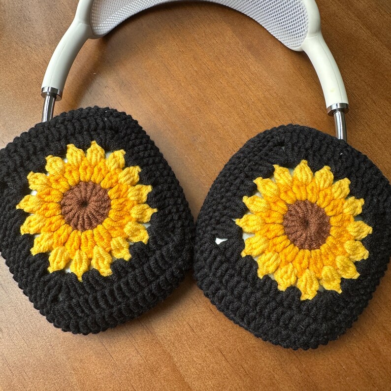 May include: Black crocheted earmuffs with sunflower designs. The sunflowers have yellow petals and brown centers.