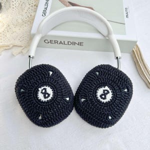 Puede incluir: Fundas para auriculares de crochet negro con un dise&ntilde;o de bola de billar blanca de 8. Las fundas est&aacute;n unidas a una diadema blanca.