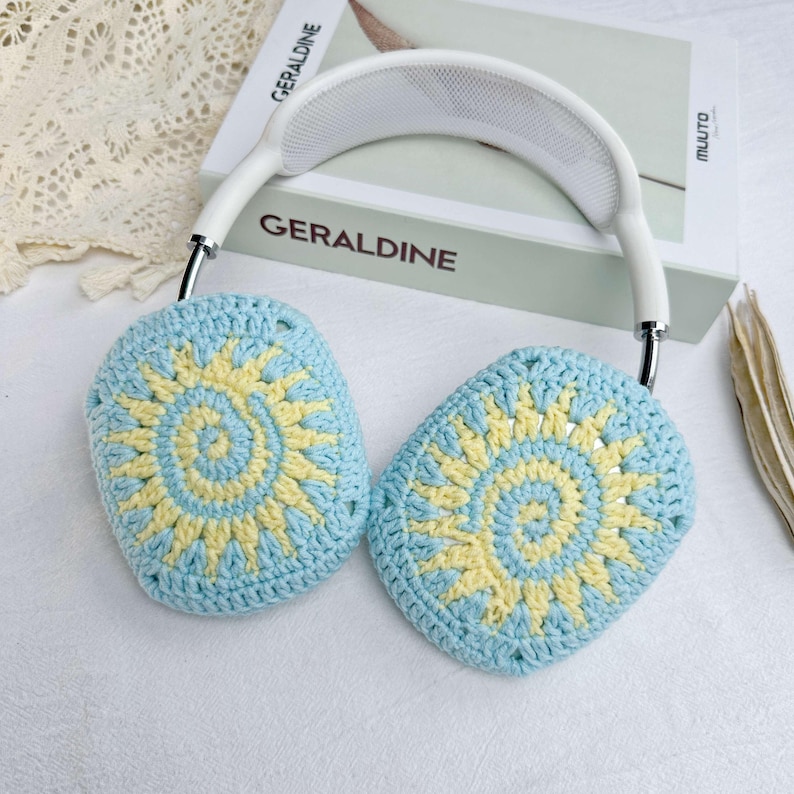 May include: White headphones with blue and yellow crocheted earmuffs featuring a sun design. The earmuffs are made with a circular pattern.