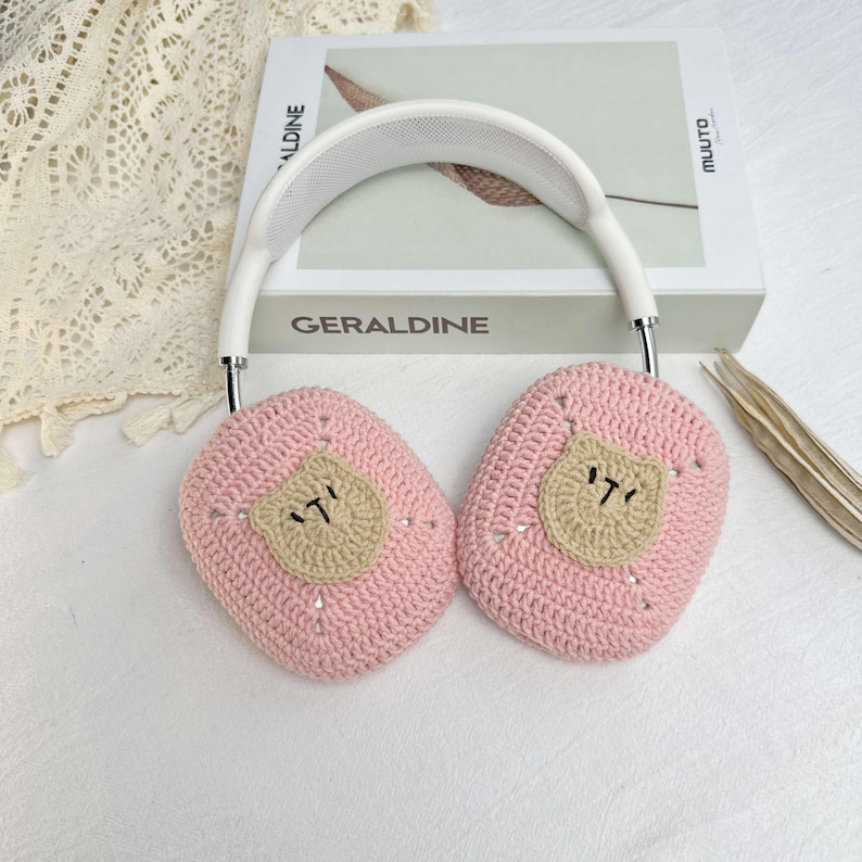 K&ouml;nnte beinhalten: Rosa gestrickte Ohrensch&uuml;tzer mit einem braunen B&auml;ren-Design. Die Ohrensch&uuml;tzer sind an einem wei&szlig;en Kopfband befestigt.
