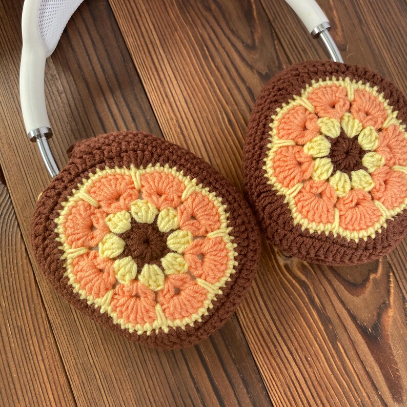 K&ouml;nnte beinhalten: Geh&auml;kelte Ohrensch&uuml;tzer in Braun, Orange und Gelb mit einem Blumenmuster. Die Ohrensch&uuml;tzer sind an einem wei&szlig;en Stirnband befestigt.