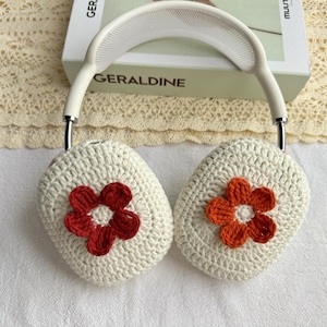 Puede incluir: Cubiertas de auriculares de crochet blanco con una flor roja y blanca en un lado y una flor naranja y blanca en el otro.