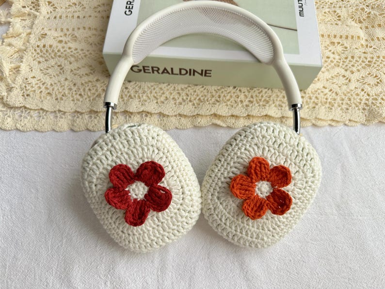 Puede incluir: Cubiertas de auriculares de crochet blanco con una flor roja y blanca en un lado y una flor naranja y blanca en el otro.