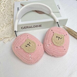 K&ouml;nnte beinhalten: Rosa gestrickte Kopfh&ouml;rerabdeckungen mit einem braunen geh&auml;kelten B&auml;renkopf auf jeder Abdeckung.