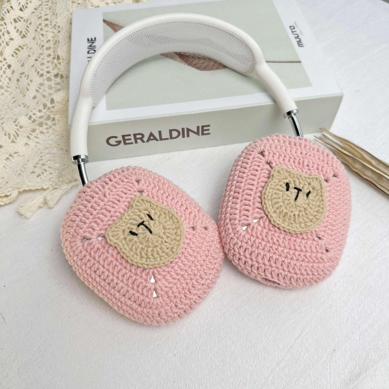 K&ouml;nnte beinhalten: Rosa gestrickte Kopfh&ouml;rerabdeckungen mit einem braunen geh&auml;kelten B&auml;renkopf auf jeder Abdeckung.
