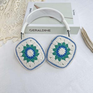 May include: White crochet earmuffs with a blue and green floral pattern. The earmuffs are attached to a white headband.