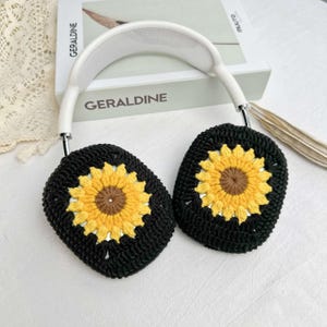 May include: White headphones with black crocheted earmuffs featuring yellow sunflower designs. The earmuffs are crocheted with a black yarn and have a yellow sunflower in the center of each earmuff.