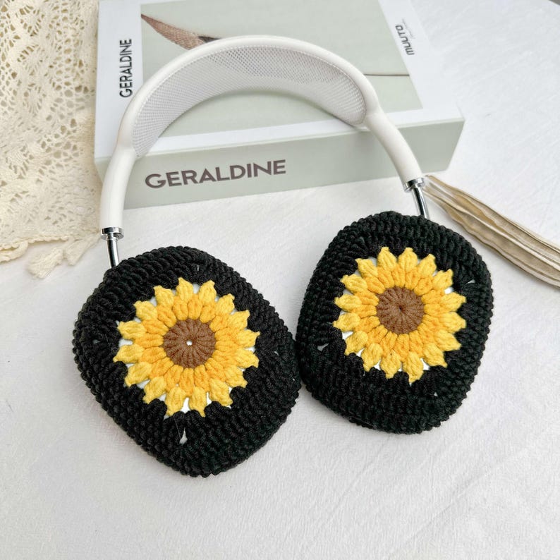 May include: White headphones with black crocheted earmuffs featuring yellow sunflower designs. The earmuffs are crocheted with a black yarn and have a yellow sunflower in the center of each earmuff.