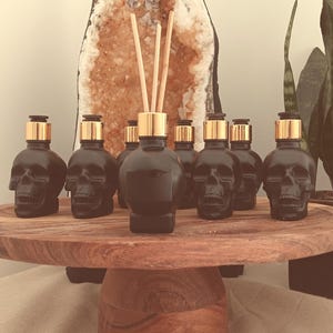 May include: Seven black glass bottles shaped like skulls with gold lids and wooden sticks. The bottles are arranged in a row on a wooden pedestal.
