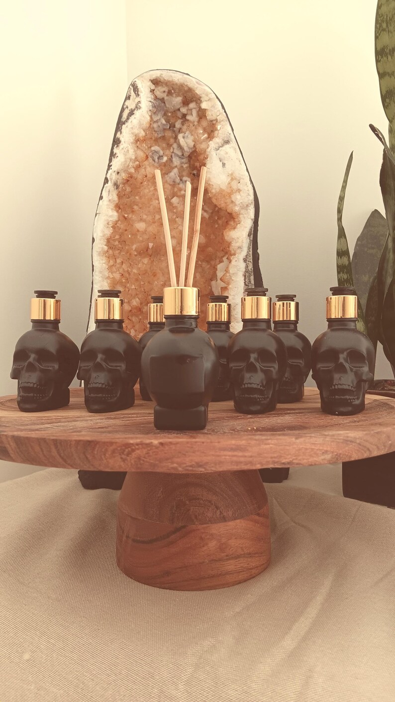 May include: Seven black glass bottles shaped like skulls with gold lids and wooden sticks. The bottles are arranged in a row on a wooden pedestal.
