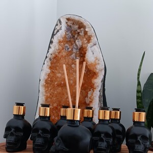 May include: A set of seven black skull-shaped glass bottles with gold lids and wooden diffuser sticks. The bottles are arranged in a semi-circle on a wooden surface. A large crystal geode is in the background.