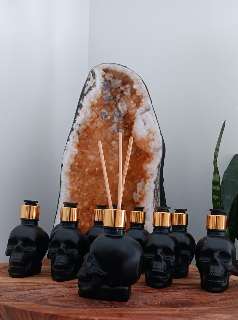 May include: A set of seven black skull-shaped glass bottles with gold lids and wooden diffuser sticks. The bottles are arranged in a semi-circle on a wooden surface. A large crystal geode is in the background.
