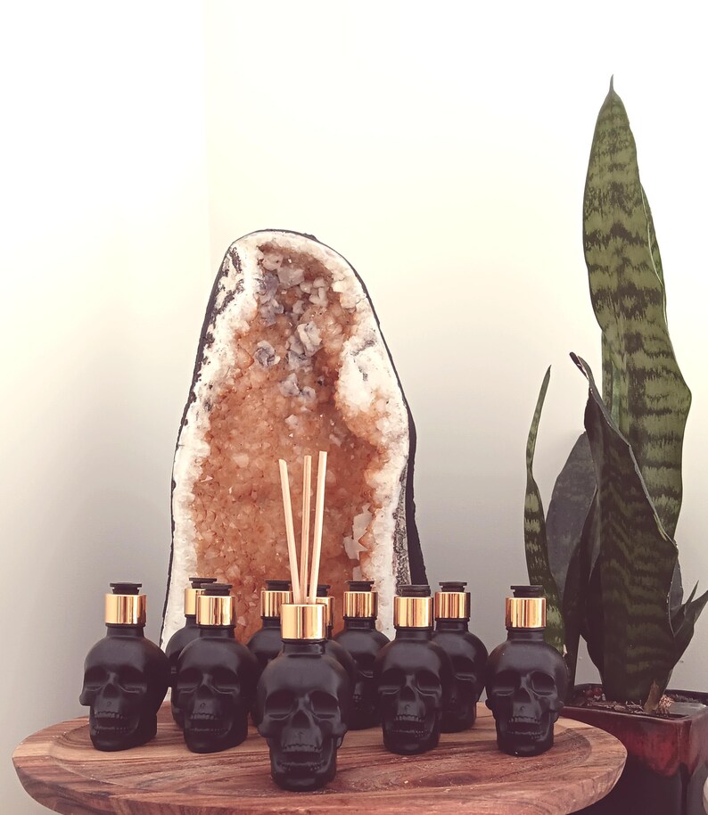 May include: A set of eight black glass skull-shaped bottles with gold lids and a set of wooden diffuser sticks. The bottles are arranged in a row on a wooden surface in front of a large crystal geode.