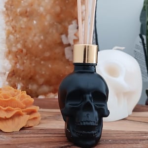 May include: Black skull shaped glass bottle with a gold lid and wooden diffuser sticks. The bottle is sitting on a wooden surface.