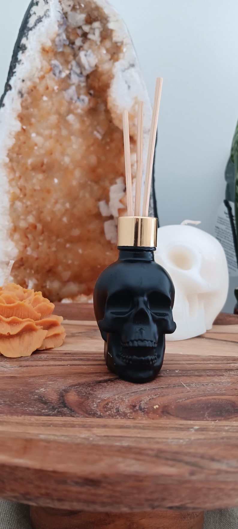 May include: Black skull shaped glass bottle with a gold lid and wooden diffuser sticks. The bottle is sitting on a wooden surface.