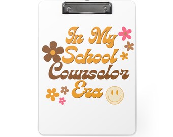 May include: White clipboard with a silver clip. The clipboard has a retro-style design with the text "In My School Counselor Era" in orange and brown lettering. There are small yellow and pink flowers and a yellow smiley face on the clipboard.