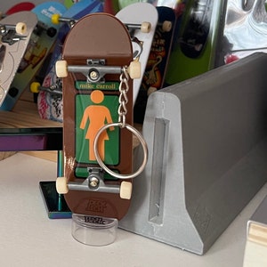 Peut inclure: Un mini skateboard marron avec un porte-clés attaché. Le skateboard a un motif représentant une silhouette féminine avec le texte "mike carroll" dessus. Le skateboard est sur un support en plastique transparent.
