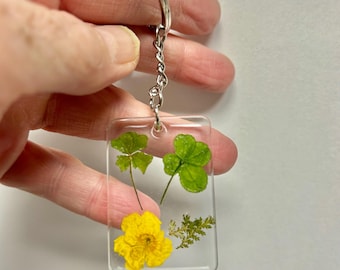 May include: A clear rectangular keychain with a silver metal ring and chain. The keychain contains a yellow flower, two green clovers, and small green leaves. The keychain is held by a hand.