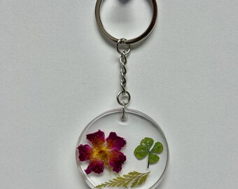 Op de afbeelding: Een ronde, transparante sleutelhanger van hars met een geperste roze bloem, een klavertjevier en een varen.