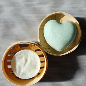May include: Two bamboo baskets each holding a soap product. One basket contains a round, white soap. The other holds a heart-shaped, pale green soap. The baskets are on a light gray wooden surface.