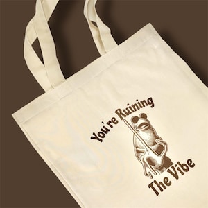 May include: Cream tote bag featuring a brown frog illustration wearing sunglasses and holding a stick. The text "You're Ruining The Vibe" is printed in brown. The bag has two cream-colored handles.