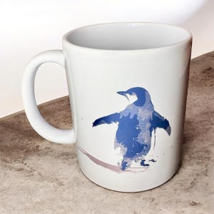 May include: White ceramic mug with a blue penguin design. The penguin is depicted with a watercolour effect and outstretched wings. The mug has a curved handle and is suitable for hot drinks. The design is simple and clean.