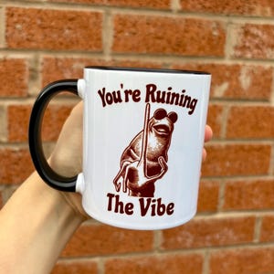 May include: White ceramic mug with a black handle and rim. The mug features a brown illustration of a frog wearing sunglasses and holding a stick, with the text "You're Ruining The Vibe" in brown.