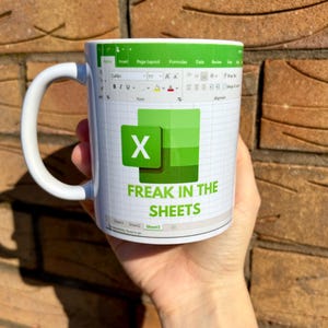 K&ouml;nnte beinhalten: Wei&szlig;e Keramiktasse mit einem gr&uuml;n-wei&szlig;en Design. Das Design zeigt eine Tabellenkalkulation mit dem Buchstaben "X" in einer gr&uuml;nen Zelle und dem Text "Freak in the Sheets".