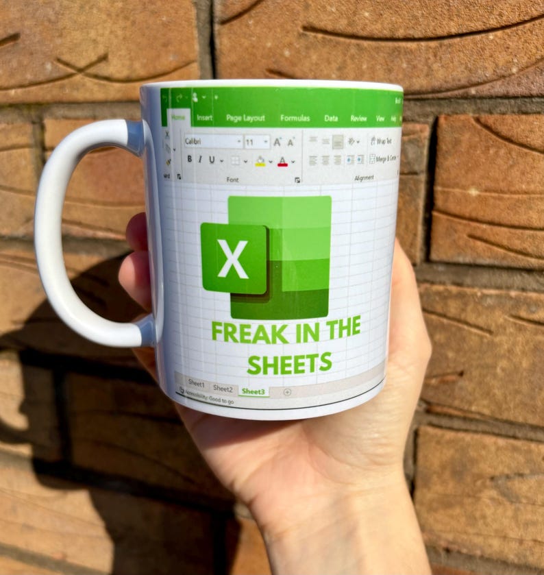 K&ouml;nnte beinhalten: Wei&szlig;e Keramiktasse mit einem gr&uuml;n-wei&szlig;en Design. Das Design zeigt eine Tabellenkalkulation mit dem Buchstaben "X" in einer gr&uuml;nen Zelle und dem Text "Freak in the Sheets".