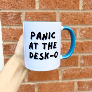 K&ouml;nnte beinhalten: Wei&szlig;er Keramikbecher mit blauem Innenraum und Henkel. Der Becher tr&auml;gt den Text "PANIC AT THE DESK-O" in schwarzen, fetten Buchstaben. Der Becher wird vor einer roten Backsteinmauer gehalten.
