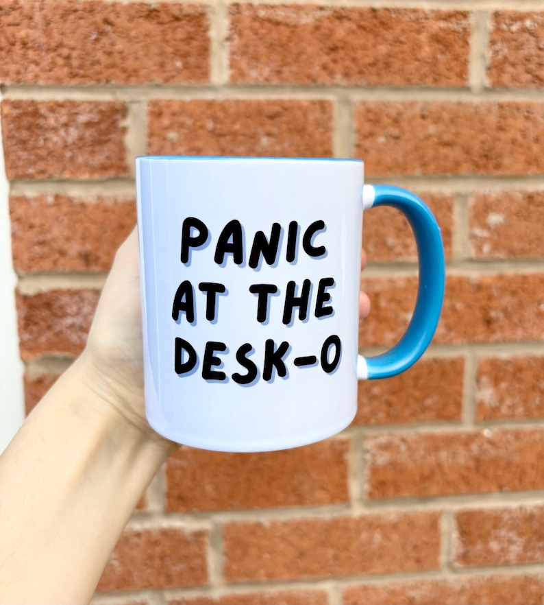 K&ouml;nnte beinhalten: Wei&szlig;er Keramikbecher mit blauem Innenraum und Henkel. Der Becher tr&auml;gt den Text "PANIC AT THE DESK-O" in schwarzen, fetten Buchstaben. Der Becher wird vor einer roten Backsteinmauer gehalten.