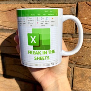 K&ouml;nnte beinhalten: Eine wei&szlig;e Kaffeetasse mit einem gr&uuml;nen Excel-Tabellenblatt-Design. Die Tasse tr&auml;gt den Text "FREAK IN THE SHEETS".