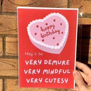 May include: A red card with a pink heart-shaped cake that says "happy birthday" in red lettering. The card says "May it be: VERY DEMURE VERY MINDFUL VERY CUTESY"