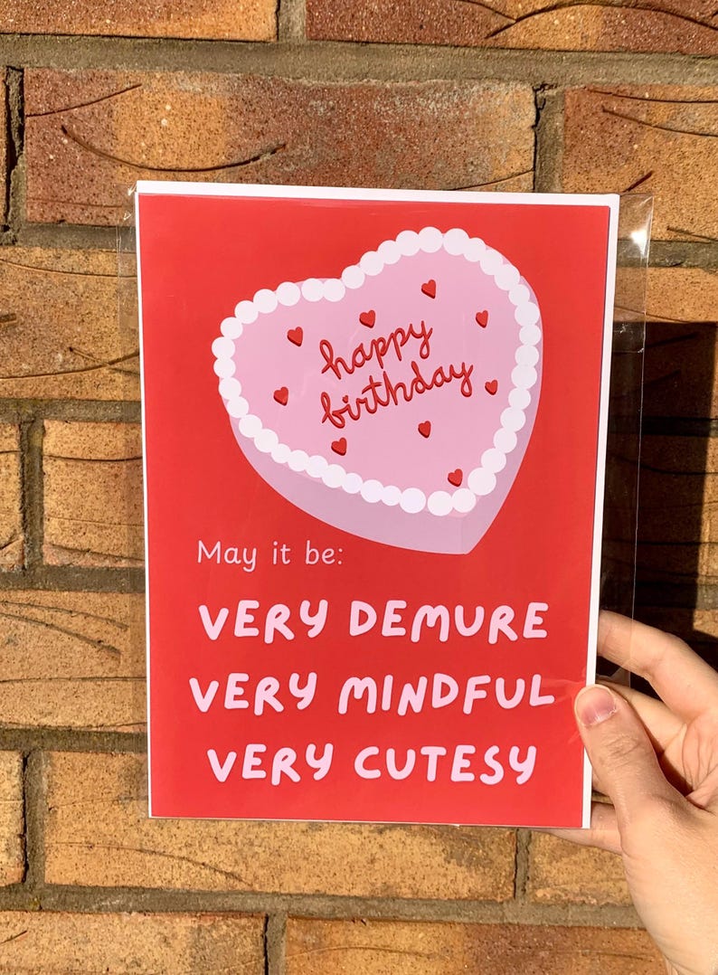 May include: A red card with a pink heart-shaped cake that says "happy birthday" in red lettering. The card says "May it be: VERY DEMURE VERY MINDFUL VERY CUTESY"