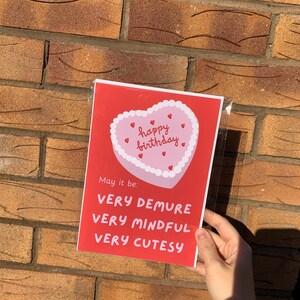 May include: Red card with a pink heart-shaped cake with white frosting and red hearts. The cake says "happy birthday". The card says "May it be: very demure, very mindful, very cutesy."