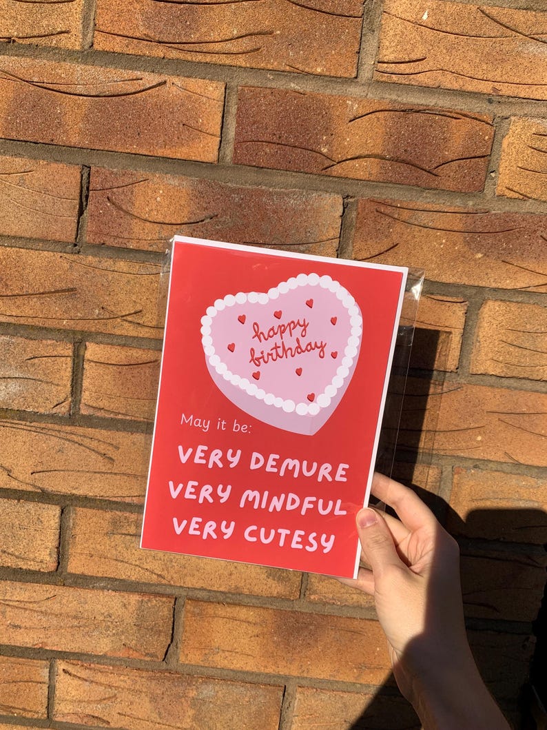 May include: Red card with a pink heart-shaped cake with white frosting and red hearts. The cake says "happy birthday". The card says "May it be: very demure, very mindful, very cutesy."