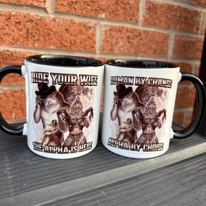 May include: Two white ceramic mugs with black handles and rims. Each mug features a werewolf design with the phrases "HIDE YOUR WIFE" and "HUMAN BY CHANCE". The text "THE ALPHA IS HERE" and "ALPHA BY CHOICE" are also printed on the mugs.