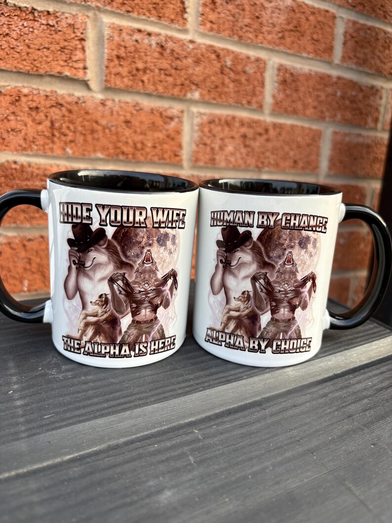 May include: Two white ceramic mugs with black handles and rims. Each mug features a werewolf design with the phrases "HIDE YOUR WIFE" and "HUMAN BY CHANCE". The text "THE ALPHA IS HERE" and "ALPHA BY CHOICE" are also printed on the mugs.