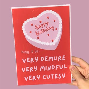 May include: Red card with a pink heart-shaped cake with white frosting and red hearts. The cake says "happy birthday". The card says "May it be: VERY DEMURE VERY MINDFUL VERY CUTESY"