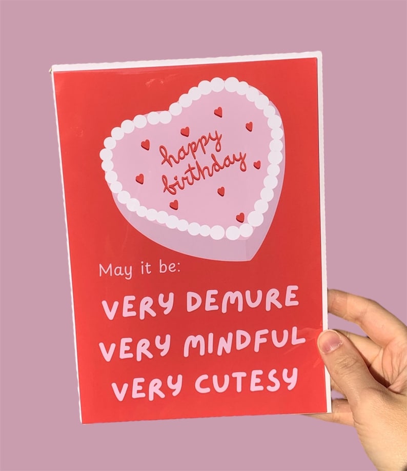 May include: Red card with a pink heart-shaped cake with white frosting and red hearts. The cake says "happy birthday". The card says "May it be: VERY DEMURE VERY MINDFUL VERY CUTESY"