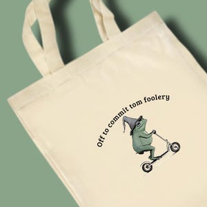 May include: Cream tote bag with a cartoon frog in a wizard hat riding a scooter. The text "Off to commit tom foolery" is above the frog. The bag has two handles. The frog is green and the hat is grey.