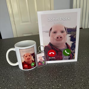 Puede incluir: Una taza blanca con una imagen de un cerdo con una camisa a cuadros rojos y negros y el texto "John Pork is calling..." con un icono de tel&eacute;fono rojo que dice "Decline" y un icono de tel&eacute;fono verde que dice "Accept". Tambi&eacute;n hay un llavero con una imagen similar del cerdo.