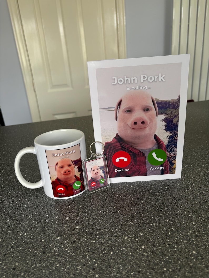 Puede incluir: Una taza blanca con una imagen de un cerdo con una camisa a cuadros rojos y negros y el texto "John Pork is calling..." con un icono de tel&eacute;fono rojo que dice "Decline" y un icono de tel&eacute;fono verde que dice "Accept". Tambi&eacute;n hay un llavero con una imagen similar del cerdo.