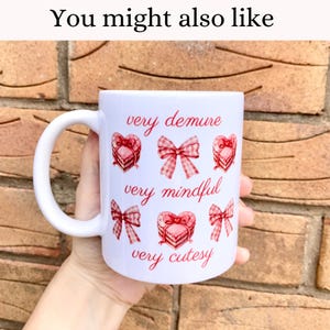May include: White ceramic mug with a pink and red design. The mug has a handle and features the text "very demure", "very mindful", and "very cutesy" with pink and red bows and hearts.