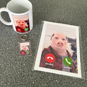 Puede incluir: Una taza blanca con una imagen de un cerdo con un tel&eacute;fono en la pantalla. El texto "John Pork is calling..." est&aacute; en la taza. Hay un llavero a juego con un cerdo y un tel&eacute;fono en la pantalla. Tambi&eacute;n hay una tarjeta con un cerdo con un tel&eacute;fono en la pantalla. El texto "John Pork is calling..." est&aacute; en la tarjeta. Hay dos botones en el tel&eacute;fono, uno rojo con la palabra "Rechazar" y uno verde con la palabra "Aceptar".