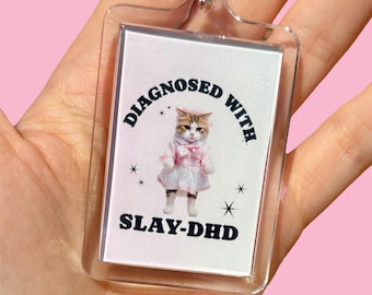 Pode incluir: Um chaveiro de acrílico transparente com fundo branco e um gato vestindo um vestido rosa. O texto no chaveiro diz "Diagnosed with Slay-DHD".
